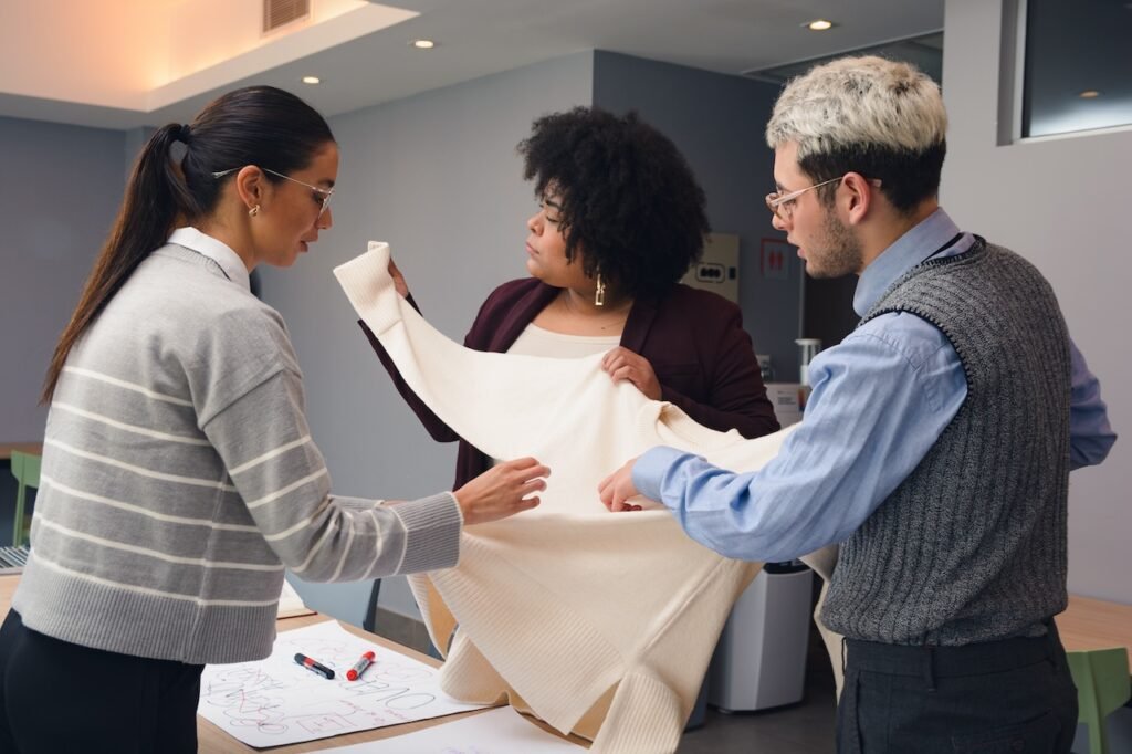 Two Women And A Man Are Standing Around A Table, One Of Them Holding And Analyzing A Piece Of Fabric