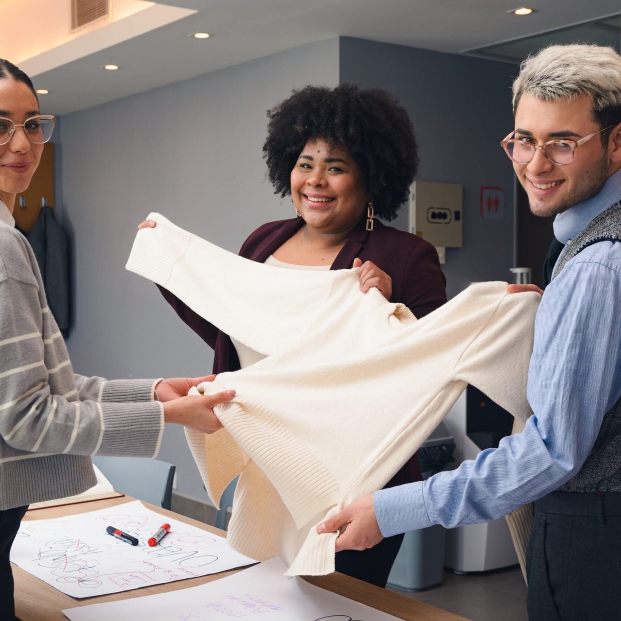 The Team of two Young Latin Women and one Man are looking at the camera smiling while working, are standing in the office happily discussing type of fabric for the clothes they are making.